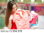 Красивая девушка с длинными волосами в красном платье делает покупки, разглядывает одежду в магазине. Стоковое фото, фотограф Вера Франц / Фотобанк Лори