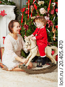 Mother sits on furry rug under Christmas tree near rocking horse on which little girl in santa suit sits. Стоковое фото, фотограф Losevsky Pavel / Фотобанк Лори