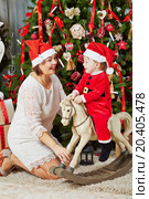Smiling mother in santa cap sits under Christmas tree near rocking horse on which little girl in santa suit sits. Стоковое фото, фотограф Losevsky Pavel / Фотобанк Лори