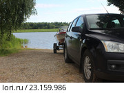 Автомобиль с катером (моторной лодкой) на прицепе (лафете, трейлере) на грунтовом слипе (спуск в воду) на фоне реки, зеленых кустов и ясного летнего неба. Стоковое фото, фотограф Илья Илмарин / Фотобанк Лори