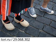 Девушка в традиционной голландской одежде и деревянных башмаках (кломпах) и мужчина в современных кедах стоят на булыжной мостовой в центре европейского города (2018 год). Редакционное фото, фотограф Николай Винокуров / Фотобанк Лори