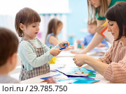 Children with scissors in hands cutting paper with teachers in class room. Group of children doing project in kindergarten. Kids play into team. Стоковое фото, фотограф Оксана Кузьмина / Фотобанк Лори