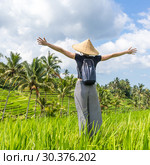 Relaxed fashionable female traveler wearing small backpack and traditional asian paddy hat, arms rised to sky, enjoying pure nature at beautiful green rice fields and terraces on Bali island. Стоковое фото, фотограф Matej Kastelic / Фотобанк Лори