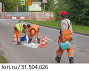 Рабочие в оранжевых спецовках льют из ведра на асфальт белую краску и разравнивают ее по заранее подготовленному трафарету. Нанесение дорожной  разметки на проезжую часть (2019 год). Редакционное фото, фотограф Наталья Николаева / Фотобанк Лори