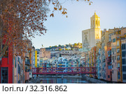 Colorful and houses and bridge Pont de Sant Agusti reflected in river Onyar, in Girona, Catalonia, Spain. Church of Sant Feliu and Saint Mary Cathedral at background in winter (2019 год). Редакционное фото, фотограф Papoyan Irina / Фотобанк Лори