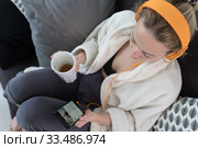 Stay at home. Social distancing. Woman at home relaxing on sofa couch drinking tea from white cup, listening to relaxing music, stay connected to friens and family via social networks on mobile. Стоковое фото, фотограф Matej Kastelic / Фотобанк Лори