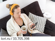 Stay at home. Social distancing. Woman at home relaxing on sofa couch drinking tea from white cup, listening to relaxing music, stay connected to friens and family via social networks on mobile. Стоковое фото, фотограф Matej Kastelic / Фотобанк Лори