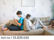 Pre-teen sister and brother gaming online with cellphones while sitting at home, children wearing safety masks, sitting back to back on sofa. Стоковое фото, фотограф Кекяляйнен Андрей / Фотобанк Лори