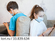 Kids playing online games with cellphones while sitting on couch indoor, boy and girl wearing medical masks. Стоковое фото, фотограф Кекяляйнен Андрей / Фотобанк Лори