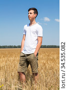 Full length portrait of teen age boy standing on agricultural field with ripe wheat, yellow meadow and blue sky, hands are in pockets of shorts pants. Стоковое фото, фотограф Кекяляйнен Андрей / Фотобанк Лори