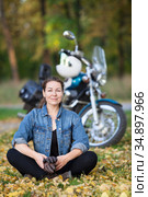 Adult biker girl sits in the lotus position in front of stylish motorcycle at autumn park, smiling happy woman on fallen leaves. Стоковое фото, фотограф Кекяляйнен Андрей / Фотобанк Лори