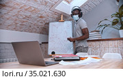 An online maths lesson - an african-american man teacher in medicine mask and big headphones standing by the board and explains the material for his students in front of the laptop. Стоковое видео, видеограф Константин Шишкин / Фотобанк Лори