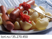 Close-up of meat sausages and boiled pasta with tomato sauce in a plate and a fork on a colorful bamboo napkin. Стоковое фото, фотограф Яна Королёва / Фотобанк Лори