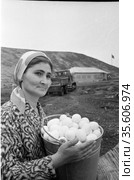 Молодая сотрудница птицефермы держит ведро с яйцами, бульдозер и самосвал рядом с домом на заднем плане, женщина занимается птицеводством, Лорино, Чукотка, 1970. Редакционное фото, фотограф Варфоломей Тетерин / Фотобанк Лори