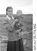 Молодая симпатичная девушка позирует стоя на поляне, она держит маленького ребёнка в руках, женщина одета в клетчатую блузку и длинную юбку, колхоз Молотова, Якутия. Редакционное фото, фотограф Варфоломей Тетерин / Фотобанк Лори