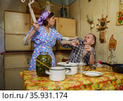 Husband drinks vodka sitting at the table in the kitchen at home, the wife swears at him and swings a rolling pin. Стоковое фото, фотограф Алексей Кузнецов / Фотобанк Лори