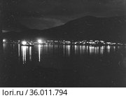 Морской пейзаж в тёмное время суток, свет от морских судов и прибрежных зданий отражается в воде, очертания холмов виднеются на противоположном берегу, Чукотка. Стоковое фото, фотограф Варфоломей Тетерин / Фотобанк Лори