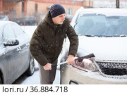 An adult Caucasian man with snow brush in hand clearing his car in winter season, swiping snow from headlight. Стоковое фото, фотограф Кекяляйнен Андрей / Фотобанк Лори