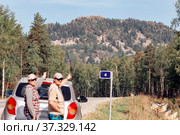 a man and a woman autotourists stand on the highway near the car and point to the high mountain Aleksandrovskaya Sopka on a sunny summer day. Стоковое фото, фотограф Акиньшин Владимир / Фотобанк Лори