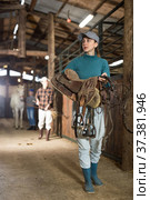 Young horsewoman walking in stable with saddle in hands. Стоковое фото, фотограф Яков Филимонов / Фотобанк Лори