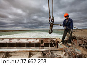 Kok-aral, Kazakhstan - May 03, 2012: Small Aral Sea Kok-aral dam. Young Asian worker lifts water shutter weighting agent. Steel rope, crane hooks. Stormy sky and turbulent water on background. Редакционное фото, фотограф Alexey Rezvykh / Фотобанк Лори