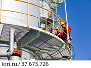 Two maintenance workers in yellow hardhats and red work wear. Distillation column of oil refinery plant on blue sky background. CNPC company (2012 год). Редакционное фото, фотограф Alexey Rezvykh / Фотобанк Лори