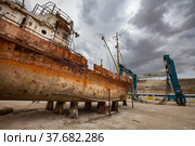 Mangystau, Kazakhstan - May 19, 2012: Old rusted ship on Caspian Sea, Bautino bay, ship-repairing yard. Grey storm cloudy sky. Marine Travelift Mobile Boat Hoist on background, right. Стоковое фото, фотограф Alexey Rezvykh / Фотобанк Лори