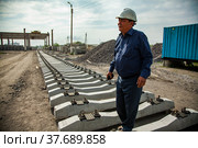 Temirtau, Kazakhstan - June 09, 2012: Concrete sleepers manufacturing plant NORD Prom NS. Asian engineer in white hardhat on outdoor sleepers warehouse. Gantry crane on background. Редакционное фото, фотограф Alexey Rezvykh / Фотобанк Лори