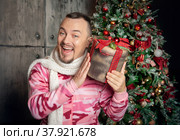 Cheerful funny happy man in pink sleepwear with Christmas present near decorated fir tree. Стоковое фото, фотограф Darkbird77 / Фотобанк Лори