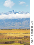 Beautiful calm rural autumn landscape with yellowed fields against the background of mountain snow-capped peaks with low clouds on a sunny day. Buryatia, Eastern Sayans, Baikal region, Tunka Valley. Стоковое фото, фотограф Виктория Катьянова / Фотобанк Лори