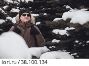 Portrait smiling brutal young man in open air among snow covered Christmas trees in winter season in forest. Tourist with glasses and backpack got lost in an unfamiliar area. Teenage crazy walks. Стоковое фото, фотограф Aleksandr Golubev / Фотобанк Лори