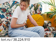 Young woman feels sick in stomach. Unhealthy female, sitting on couch and holding her stomach, feeling uncomfortable. Concept of health problems, food poisoning, gastritis. Selective focus on hands. Стоковое фото, фотограф Aleksandr Golubev / Фотобанк Лори