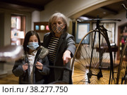 Tween schoolgirl and elderly female tutor in face masks viewing vintage bicycle in museum. Стоковое фото, фотограф Яков Филимонов / Фотобанк Лори