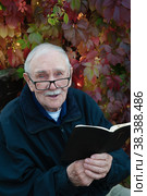 cheerful gray-haired old man with glasses is reading the Bible. Portrait of a smiling old man of 88 years on the background of beautiful autumn leaves. Стоковое фото, фотограф Оксана Медведева / Фотобанк Лори