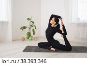 Женщина в черной спортивной одежде занимается йогой, делает упражнение Эка Пада Раджакапотасана, поза голубя, тренируется одна сидя на коврике в комнате у окна. Стоковое фото, фотограф Сергей Чайко / Фотобанк Лори