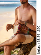 Bearded young man drummer, playing rhythm on djembe drum musical instrument on tropical sandy beach at ocean background. Travel vacation and music relaxation concept. Selective focus. Copy text space. Стоковое фото, фотограф Aleksandr Golubev / Фотобанк Лори