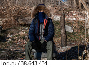 A grown man wearing a mask, wearing a hooded jacket, and wearing gloved military pants sits and looks into the camera. Стоковое фото, фотограф Владимир Арсентьев / Фотобанк Лори