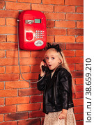 Portrait little emotional girl in leather jacket calling old red payphone at brick wall background, looking at camera. Emotion cute lady talking telephone. Communication lifestyle concept. Copy space. Стоковое фото, фотограф Aleksandr Golubev / Фотобанк Лори