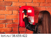 Rear view little girl in leather jacket takes handset old red payphone at brick wall backdrop. Little brunette lady model with telephone. Communication concept. Copy space for advertising text. Стоковое фото, фотограф Aleksandr Golubev / Фотобанк Лори