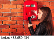 Funny little girl in leather jacket holding handset old red payphone at brick wall backdrop, looking away. Little serious lady model with phone. Communication concept. Copy space for advertising text. Стоковое фото, фотограф Aleksandr Golubev / Фотобанк Лори