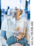 Thoughtful young mother looking trough window holding his infant baby boy child while waiting to board an airplane at airport terminal departure gates. Travel with baby concept. Стоковое фото, фотограф Matej Kastelic / Фотобанк Лори