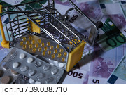 medicines and pills are lying in a small shopping cart against the background of money euro dollars 100 500. pills are poured out of the cart for money the problem of the cost of medicines expensive m. Стоковое фото, фотограф Meshkov Sergey / Фотобанк Лори