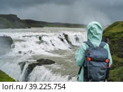 Woman with backpack and green jacket standing in front of Gullfoss powerful famous waterfall in Iceland. Tourist attraction who follow Golden Circle route of Iceland. Стоковое фото, фотограф Igor Tichonow / Фотобанк Лори
