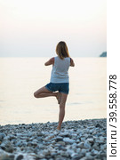 A woman leading a healthy lifestyle and practicing yoga, performs vrikshasana exercise on the seashore, tree pose, stands at sunset against the background of the sea. Стоковое фото, фотограф Сергей Чайко / Фотобанк Лори