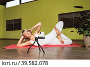 An attractive blonde woman, sports physique conducts a stream for her audience, lies on a mat and performs a lotus pose, conducts a workout online in the studio. Стоковое фото, фотограф Сергей Чайко / Фотобанк Лори
