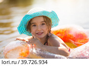 Girl in a hat stands on the river bank with a transparent inflatable circle in the shape of a heart with orange feathers inside. Beach holidays, swimming, tanning, sunscreens. Стоковое фото, фотограф Ольга Симонова / Фотобанк Лори