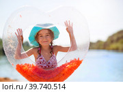 Girl in a hat stands on the river bank with a transparent inflatable circle in the shape of a heart with orange feathers inside. Beach holidays, swimming, tanning, sunscreens. Стоковое фото, фотограф Ольга Симонова / Фотобанк Лори
