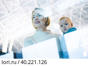 Thoughtful young mother looking trough window holding his infant baby boy child while waiting to board an airplane at airport terminal departure gates. Travel with baby concept. Стоковое фото, фотограф Matej Kastelic / Фотобанк Лори