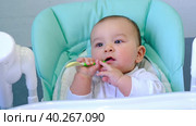 A hungry baby is gnawing on a plastic spoon at the table on a high chair. Teething, whims, itchy gums, introduction of complementary foods. Стоковое видео, видеограф Ольга Симонова / Фотобанк Лори