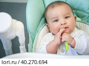 A hungry baby is gnawing on a plastic spoon at the table on a high chair. Teething, whims, itchy gums, introduction of complementary foods. Стоковое фото, фотограф Ольга Симонова / Фотобанк Лори